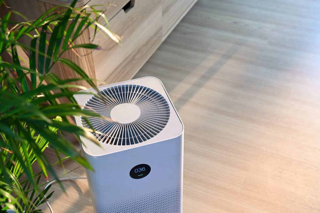 Air purifier on light wooden floor in comfortable home, with a potted house plant to the left.