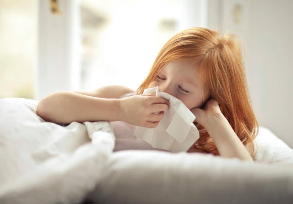 Young girl sneezing into a tissue, laying in bed at home.